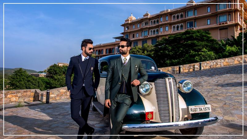 Two stylish men dressed in three-piece formal suits and sunglasses pose confidently beside a vintage car on a stone driveway. Behind them is a grand, multi-storeyed heritage-style hotel surrounded by greenery and hills, under a clear blue sky