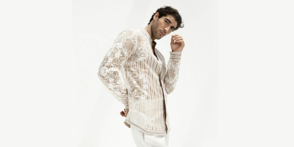 A man posing against a plain white background, wearing a stylish sheer lace shirt with intricate floral embroidery over a beige and white striped base, paired with white trousers. The outfit blends elegance and bold fashion elements.