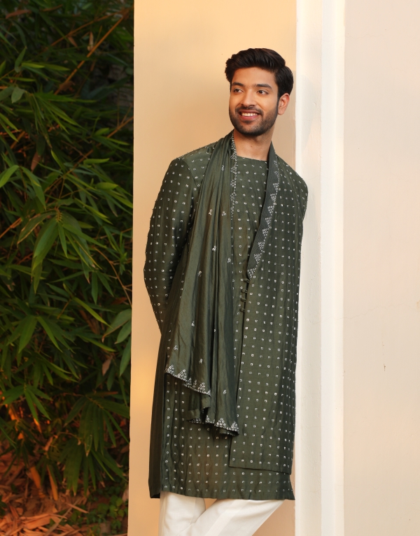 A smiling man with neatly styled hair wearing a dark green embroidered kurta with a matching dupatta and white trousers, leaning against a cream-colored wall with lush green bamboo plants in the background