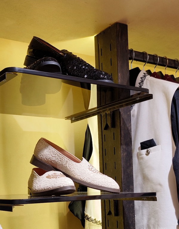 Luxury ethnic footwear displayed on glass shelves, featuring black studded formal shoes and white embroidered loafers, with elegant sherwanis and jackets hanging in the background