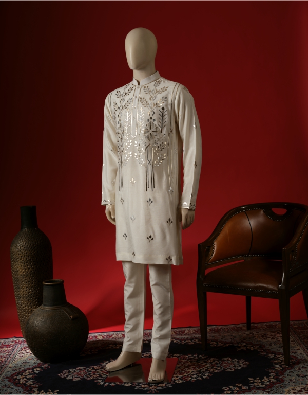 Mannequin dressed in an elegant white embroidered kurta set with intricate silver detailing, paired with matching churidar pants, set against a bold red background with traditional decor elements