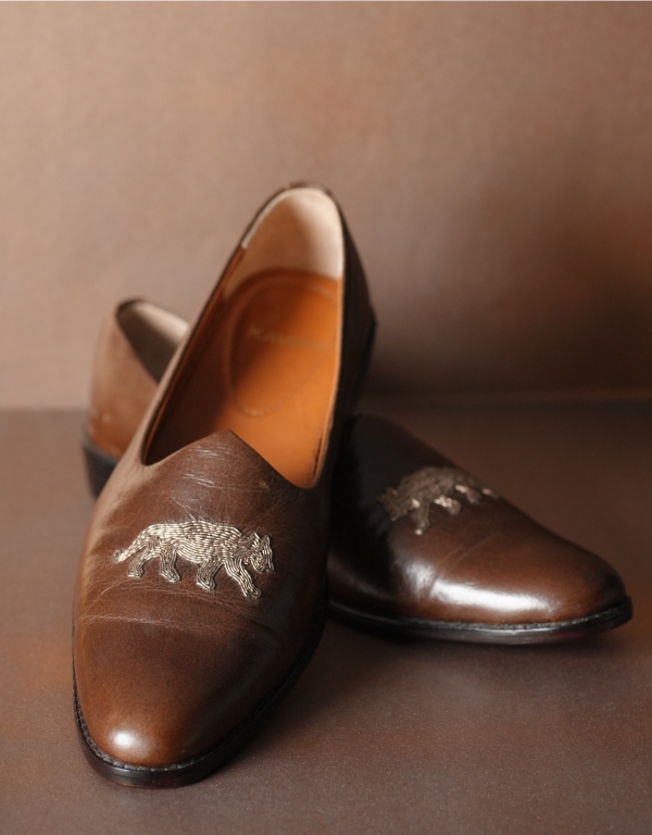 Pair of brown leather traditional Indian jutti shoes featuring an embroidered elephant motif on the front, displayed against a matching brown background.