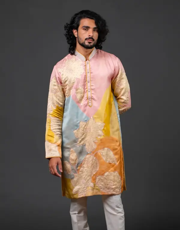 Man wearing a multicolored silk kurta with abstract floral embroidery in pastel shades of pink, blue, yellow, and orange, paired with white churidar pants, standing against a plain dark background
