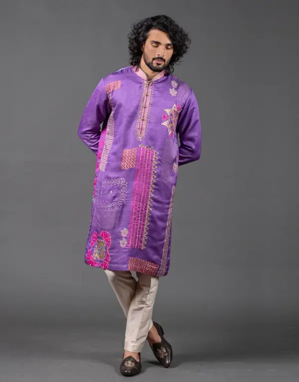 Man wearing the Satin Silk Statement Kurta Set from The HUB Menswear, featuring a vibrant purple satin silk kurta with bold, multicolored embroidered patterns and geometric motifs, paired with beige churidar pants and dark brown embroidered loafers, set against a plain grey background.